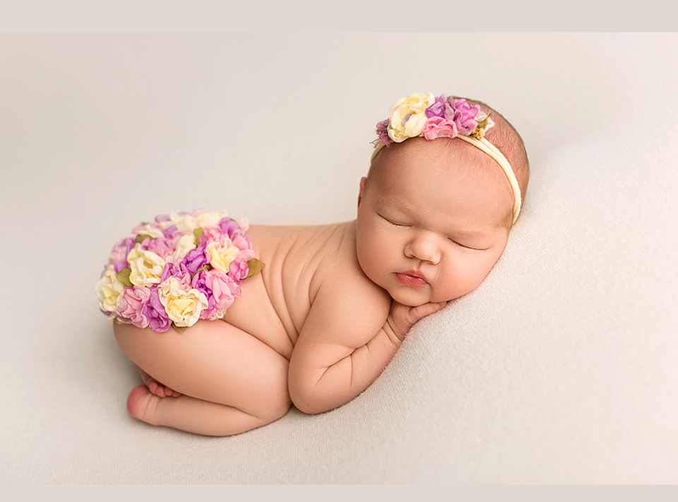 Sleeping newborn wearing floral accessories on white backdrop.