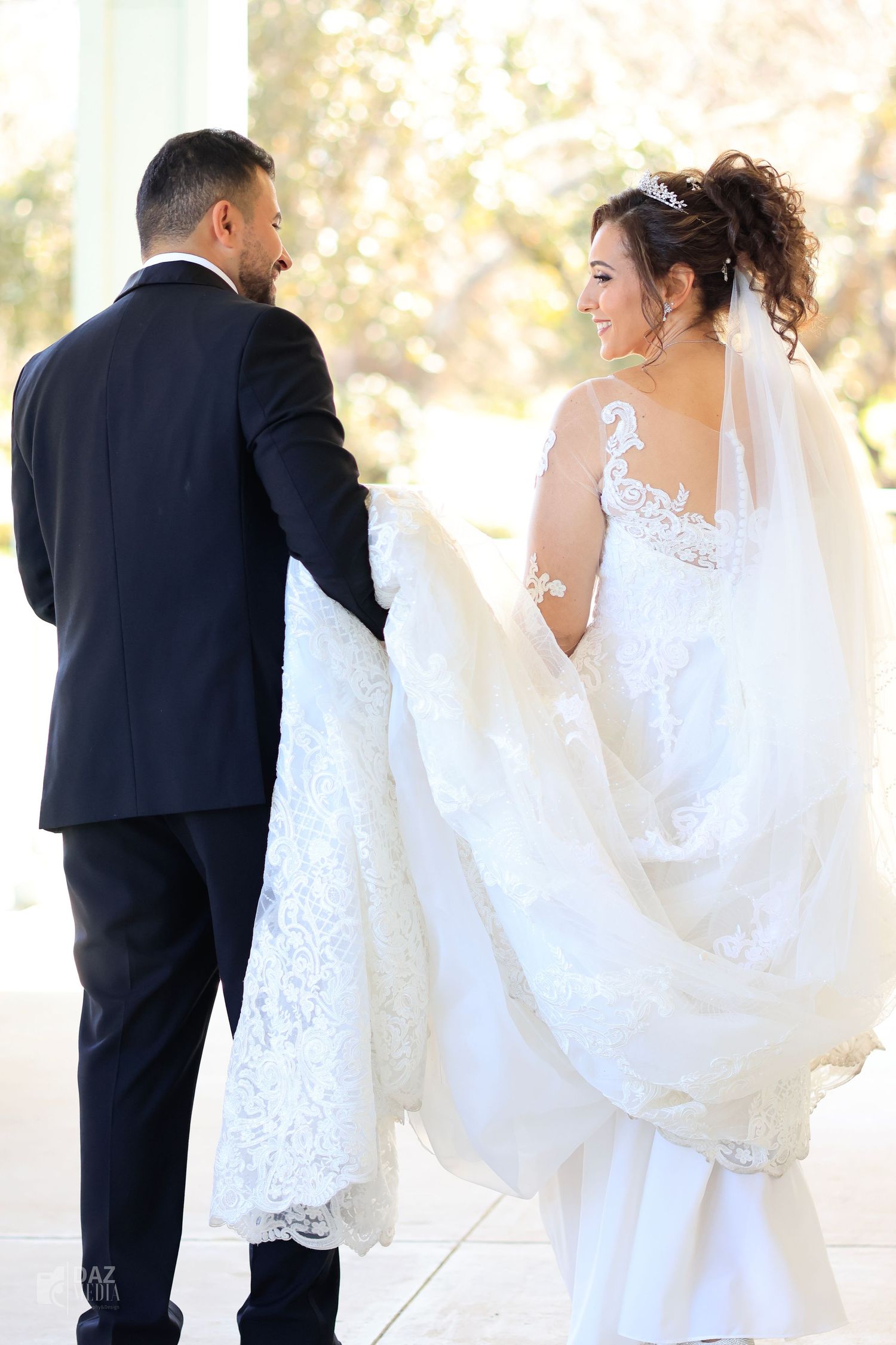 A bride and groom share a joyful moment on their wedding day, with her beautiful lace dress flowing in soft light.