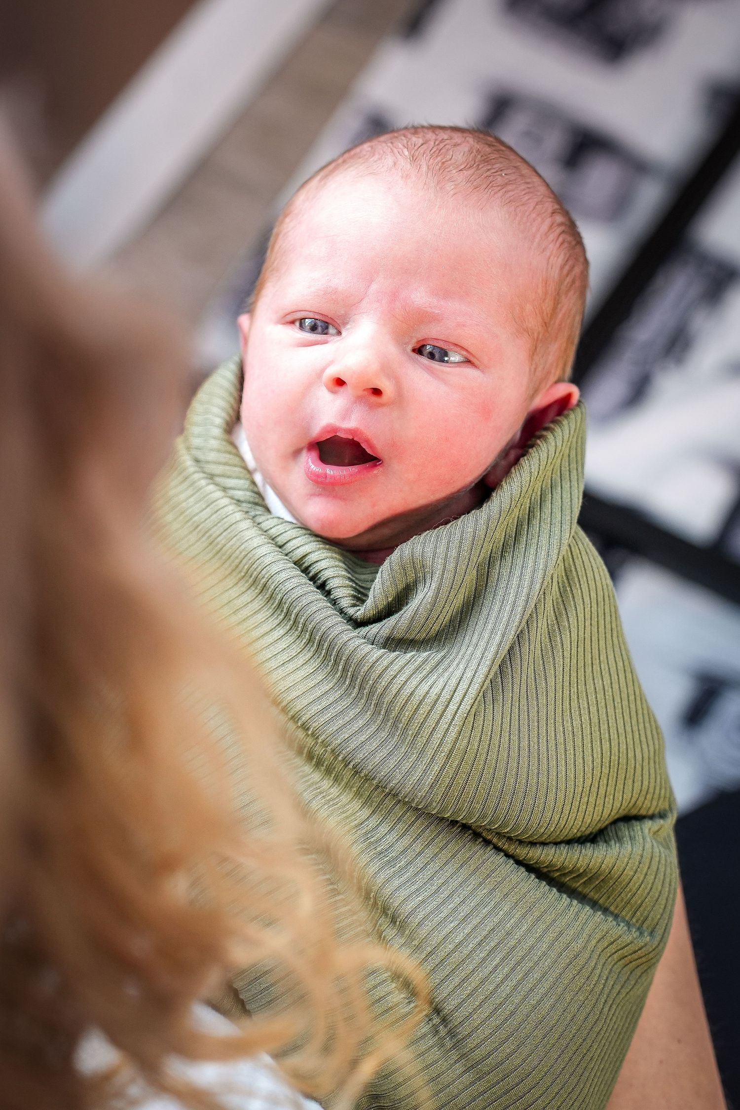 A newborn baby wrapped in a sage green knit blanket making an expressive face while looking at the camera.
