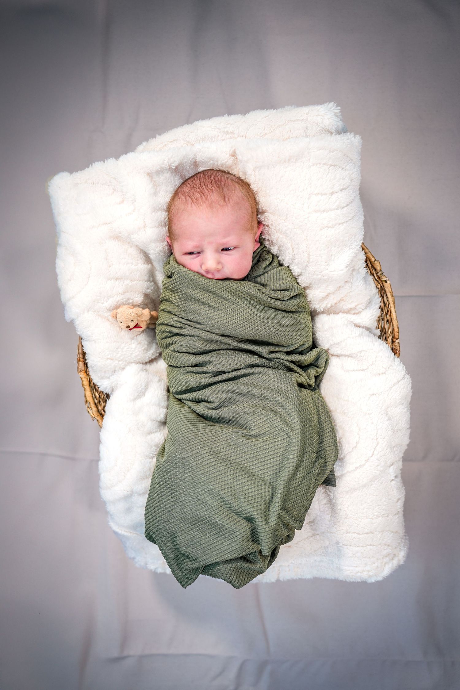 A sleeping newborn wrapped in an olive green swaddle blanket lies on a plush white cushion in a woven basket.