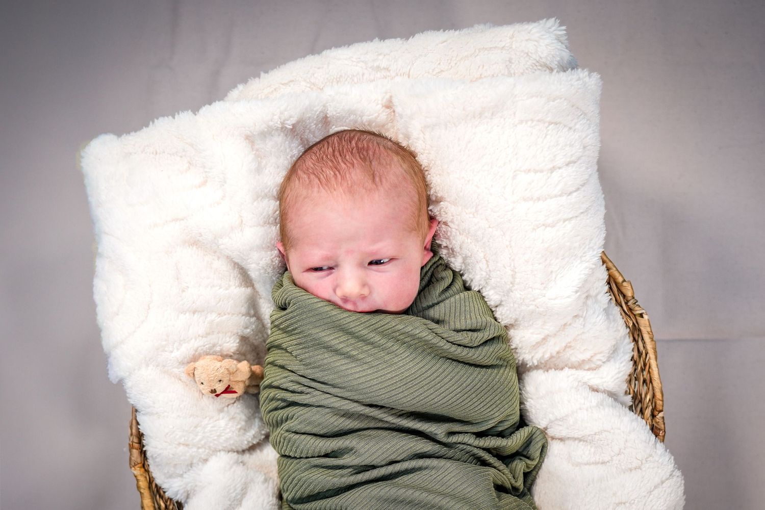 A newborn baby swaddled in a green wrap rests on a white pillow in a rustic woven basket.