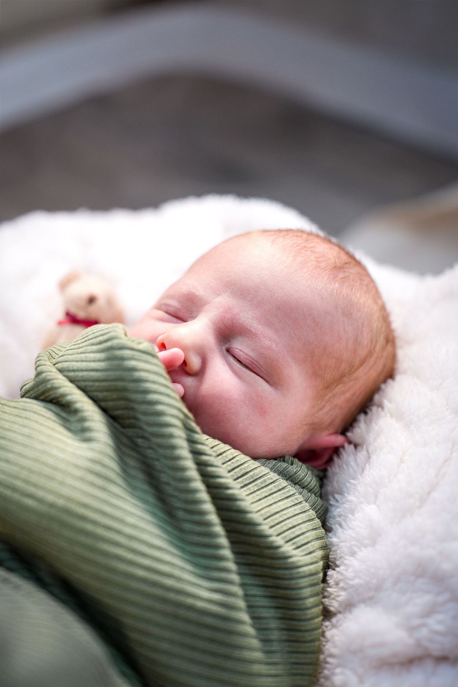 A peacefully sleeping baby wrapped in a green ribbed sweater rests on soft white bedding.