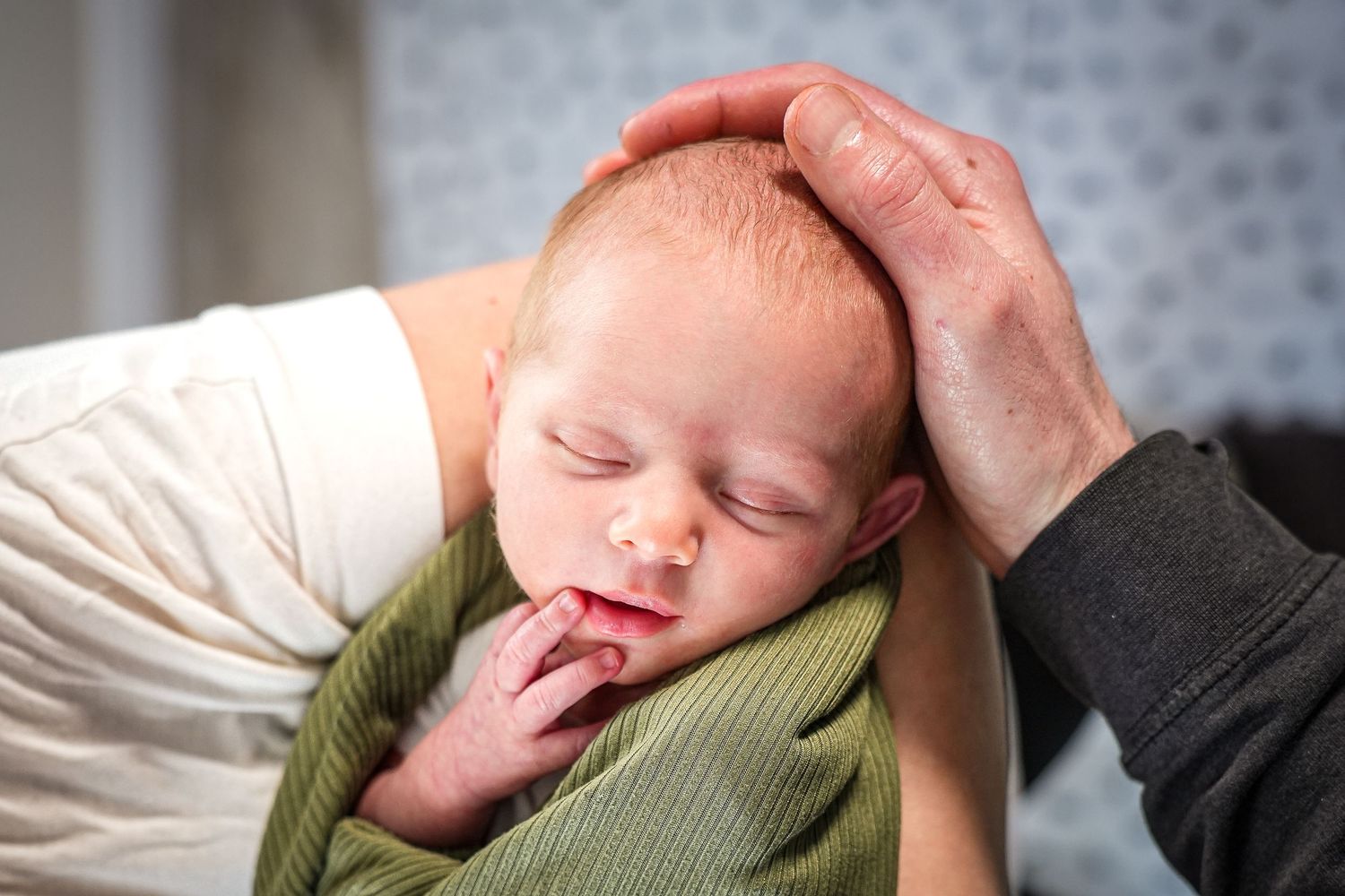 A sleeping newborn baby wrapped in a green blanket with a hand gently cradling their head.