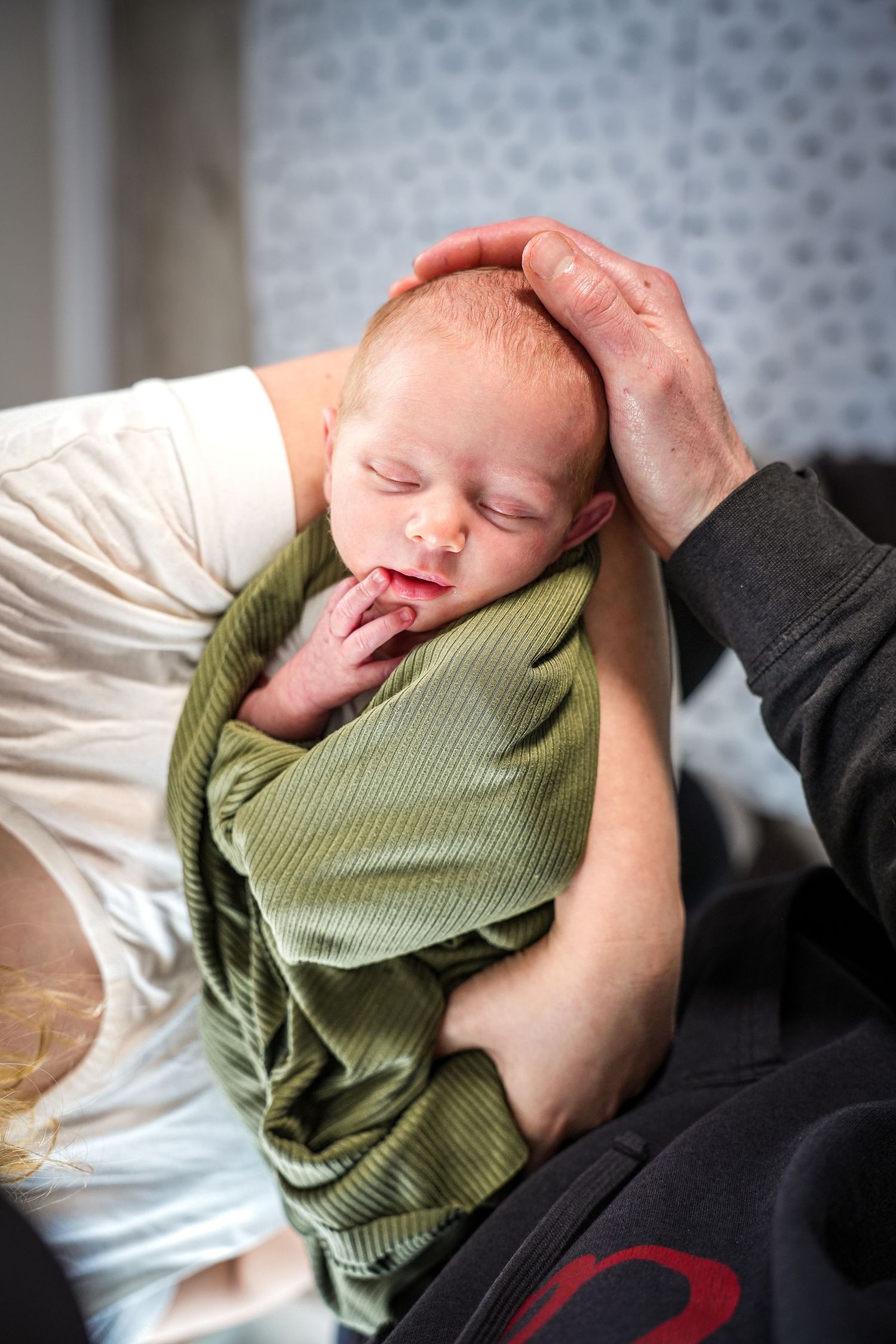 A peaceful sleeping newborn swaddled in green knit fabric being cradled by gentle hands against a soft background.