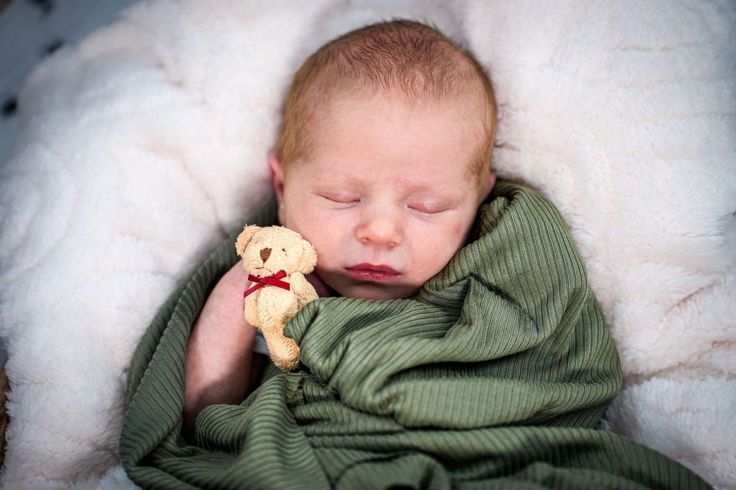 Sleeping baby in green clothing cuddles small teddy bear while resting on white blanket.