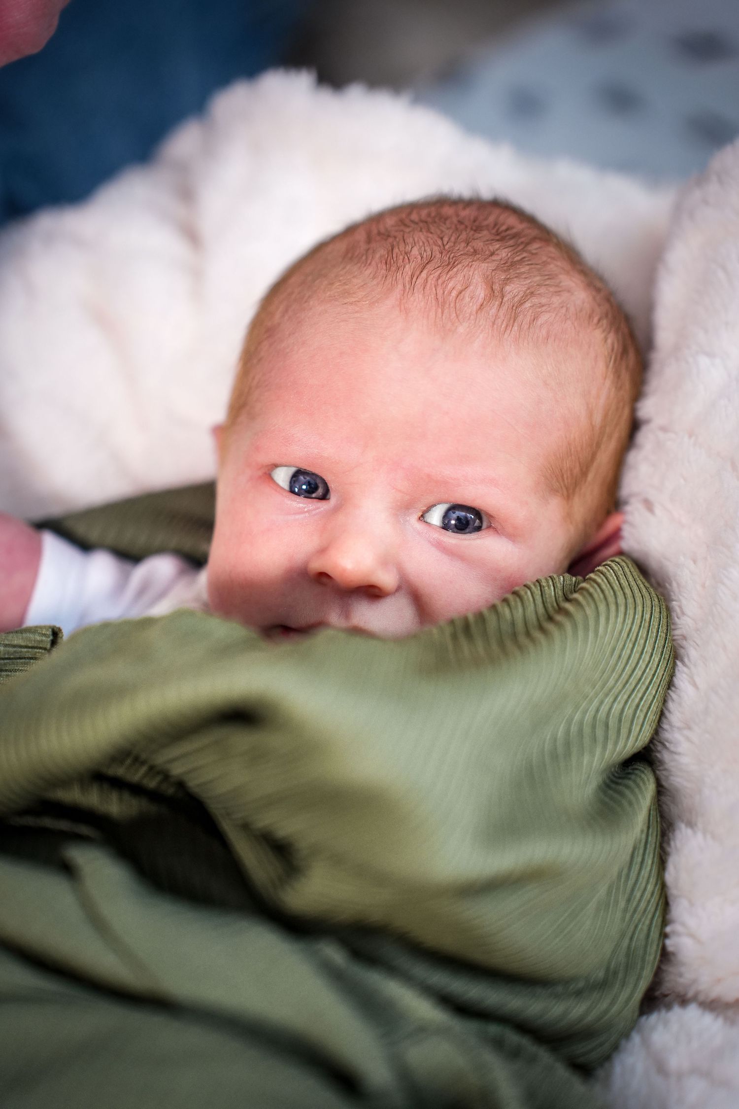 A baby wrapped in a green blanket looks up with bright blue eyes while lying on white bedding.
