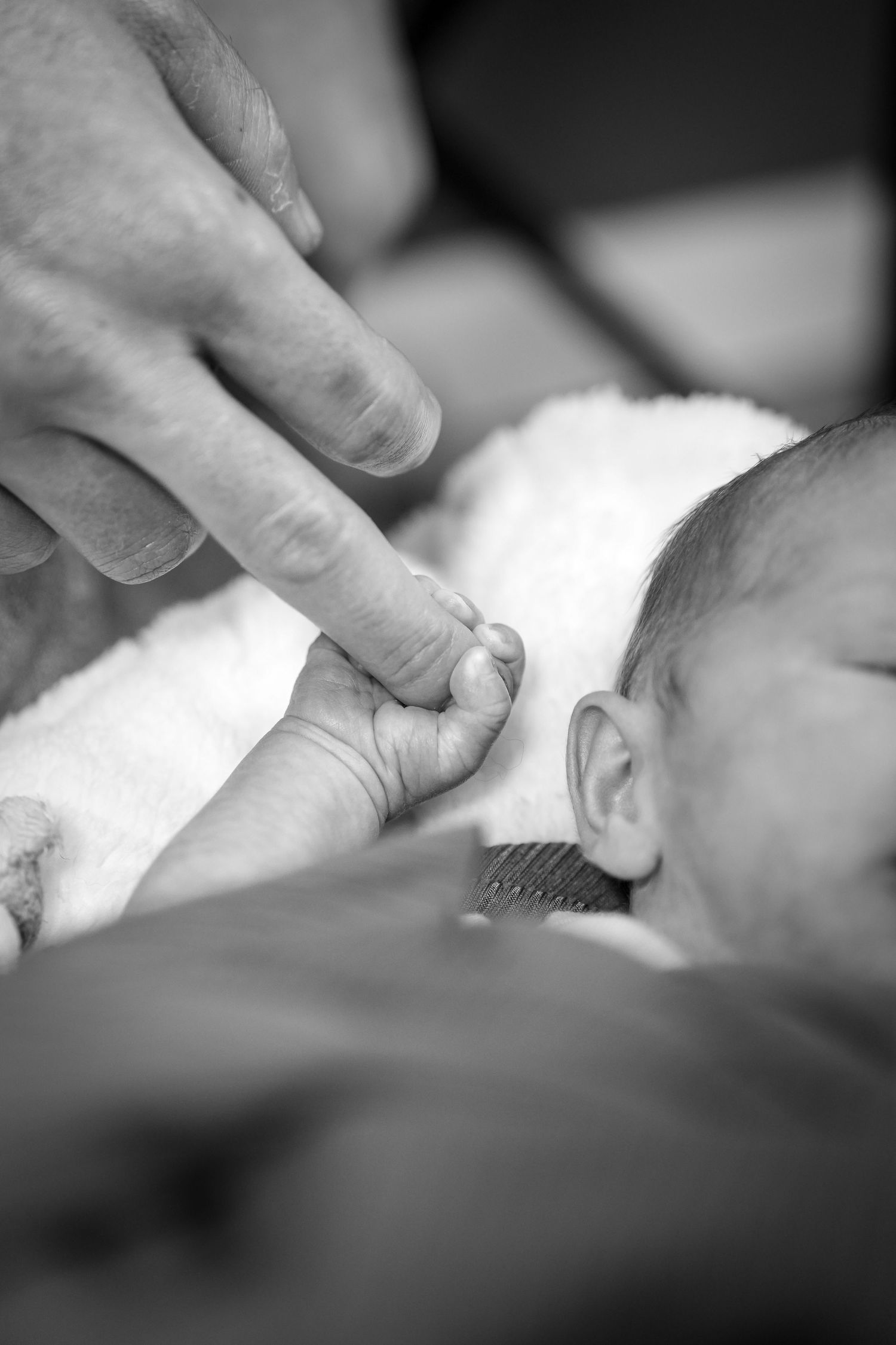 A tender moment captures a tiny infant's hand grasping an adult finger while wrapped in a white blanket, shown in black and white.
