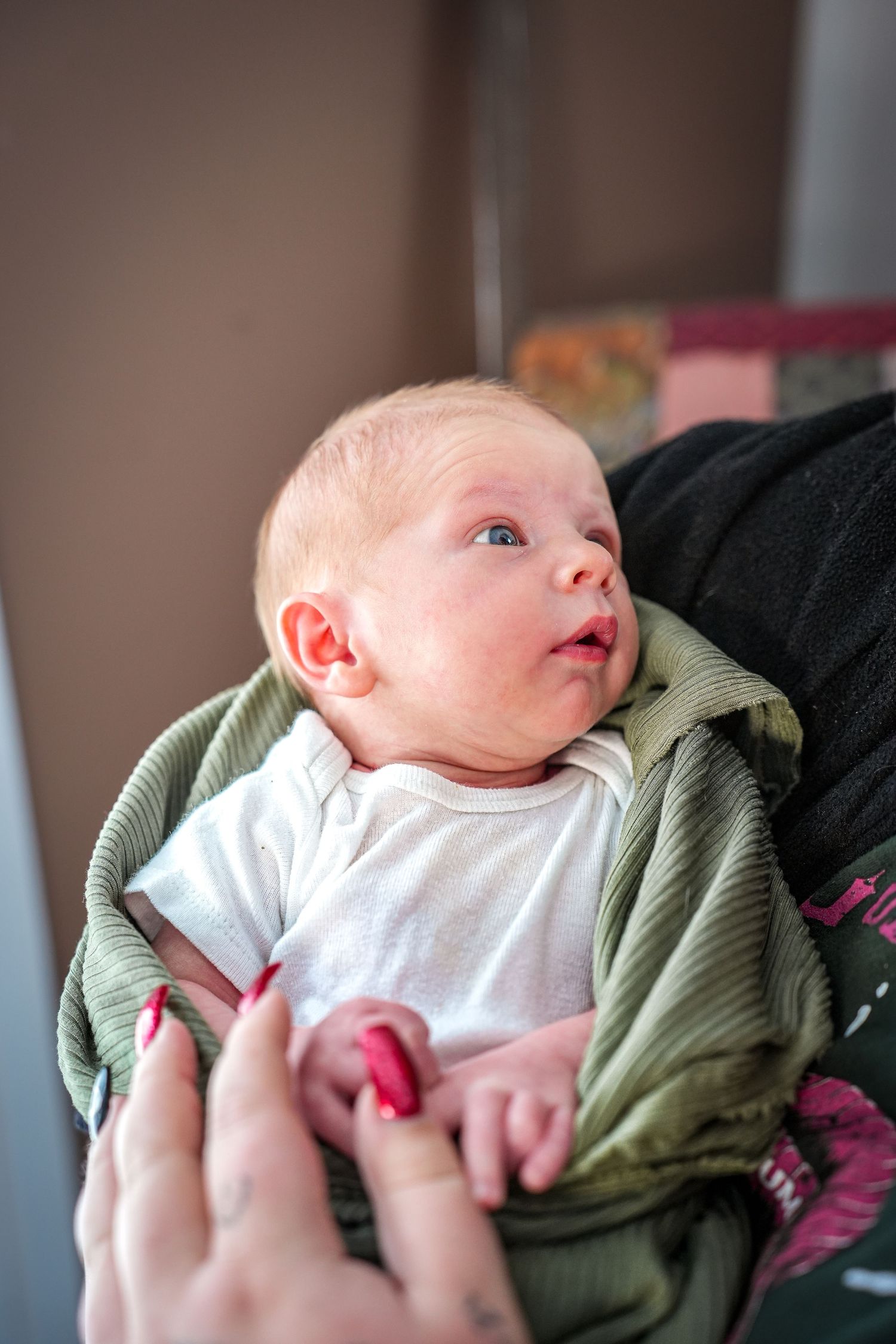 Newborn baby wrapped in green blanket looking up with curious expression while being held by caregiver.