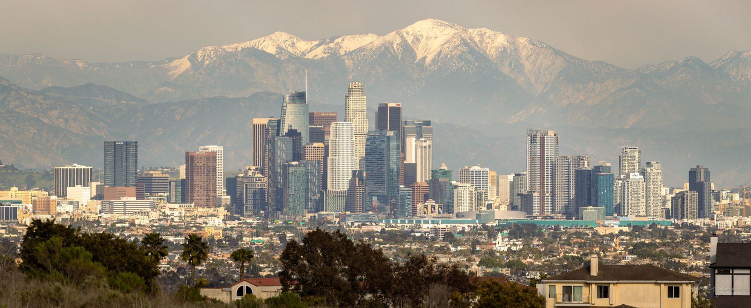 Snow-capped mountains and city skyline create a dramatic panoramic landscape at sunset.