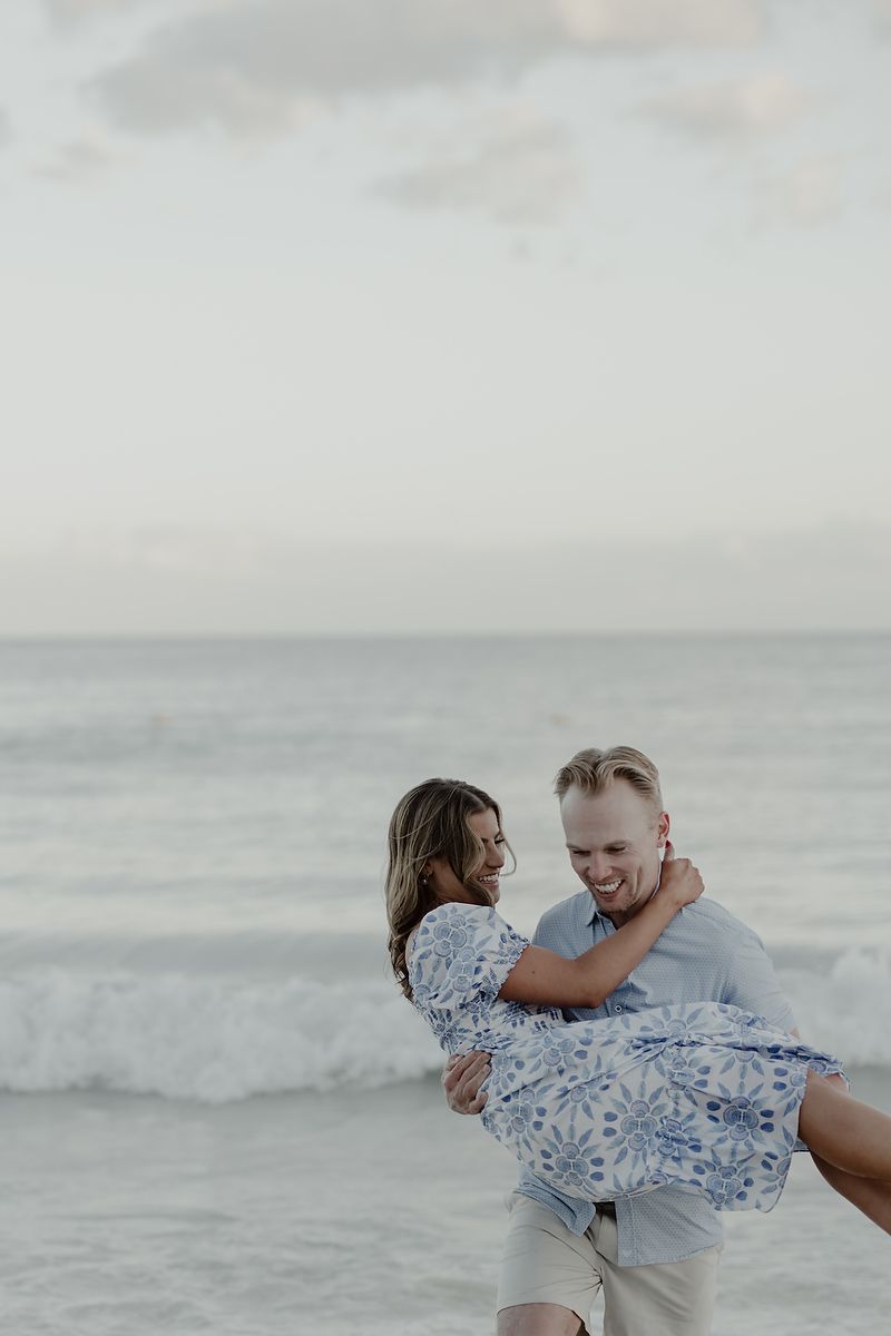 Playful engagement photoshoot with couple laughing and embracing by the turquoise sea in Riviera Maya.