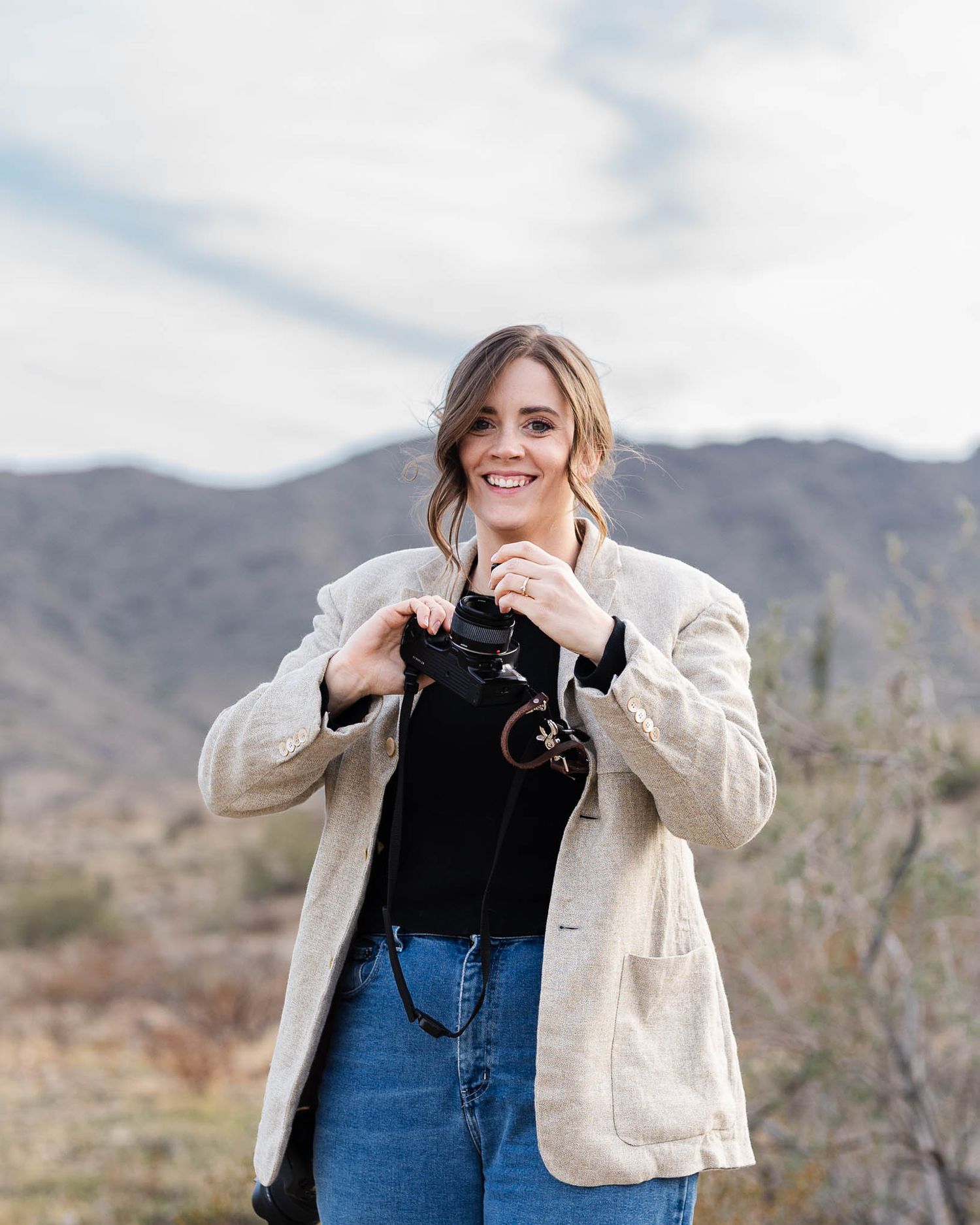 Photographer in cream jacket and jeans holds camera while smiling in front of mountain landscape.