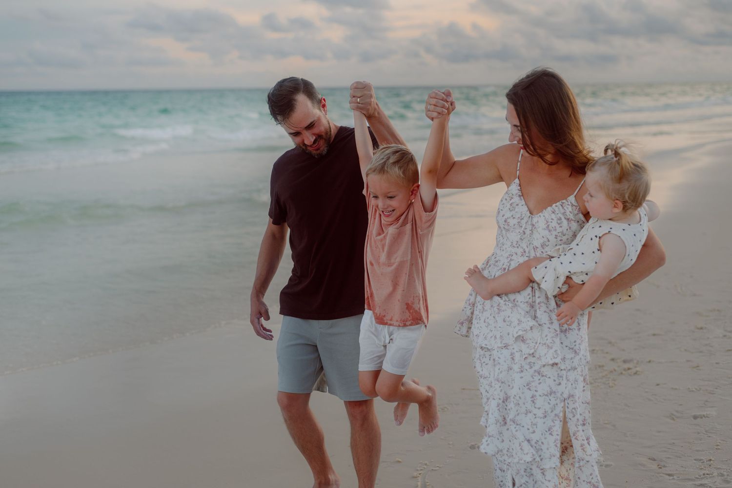 A family plays together on the beach during sunset, lifting their children in the air.