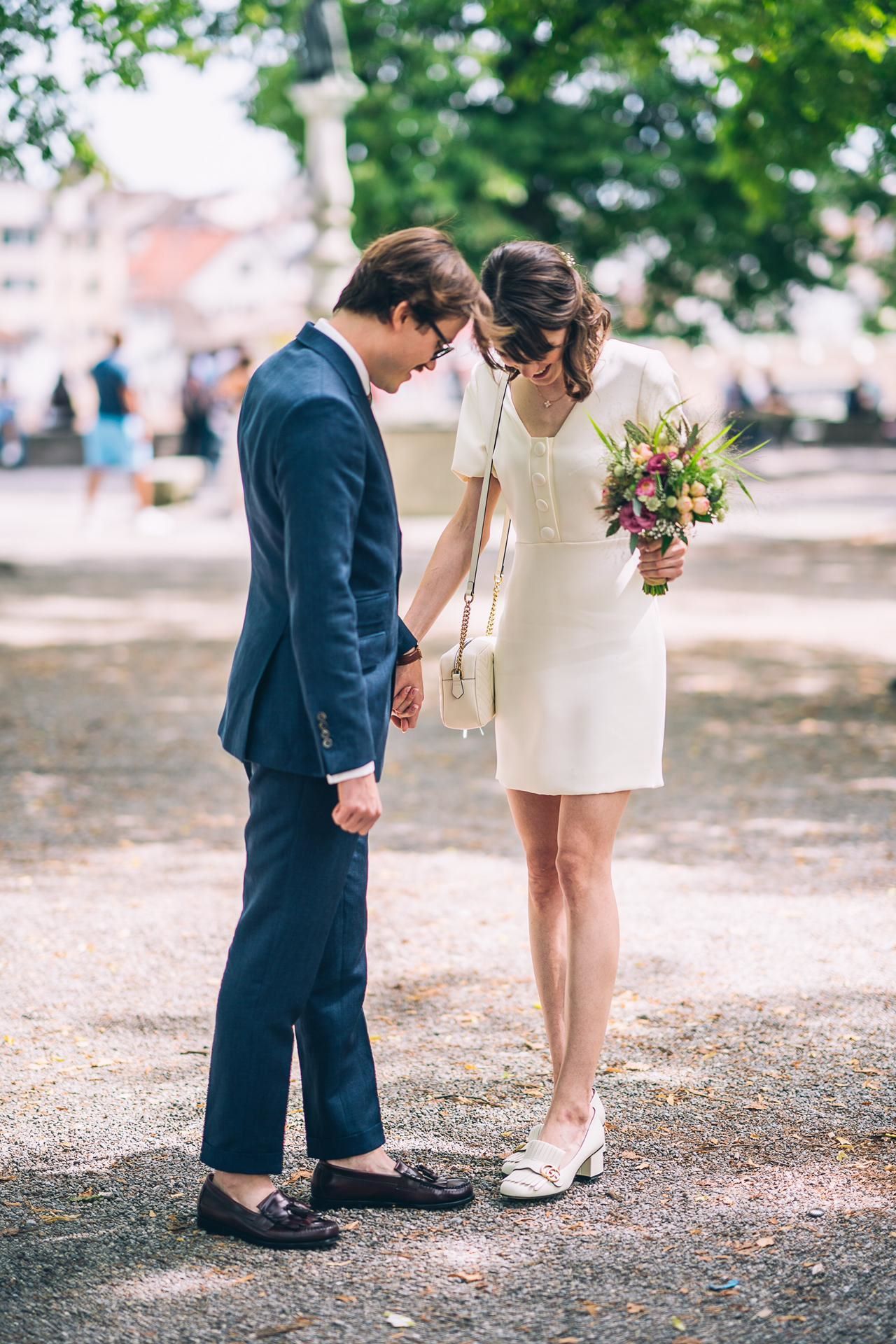 Brautpaar in einem Park. Die Braut trägt ein kurzes Kleid und der Bräutigam einen blauen Anzug. Die Braut hat einen Brautstrauss in der Hand