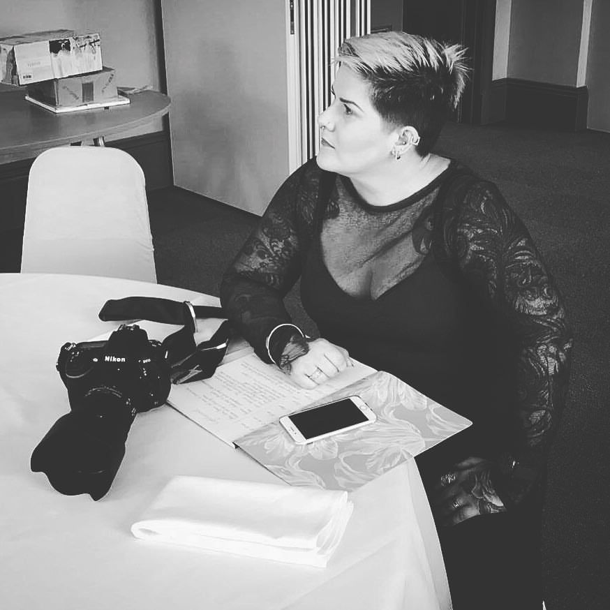 A black and white photo showing someone at a desk with a camera and notebook in a lace top looking to the side.