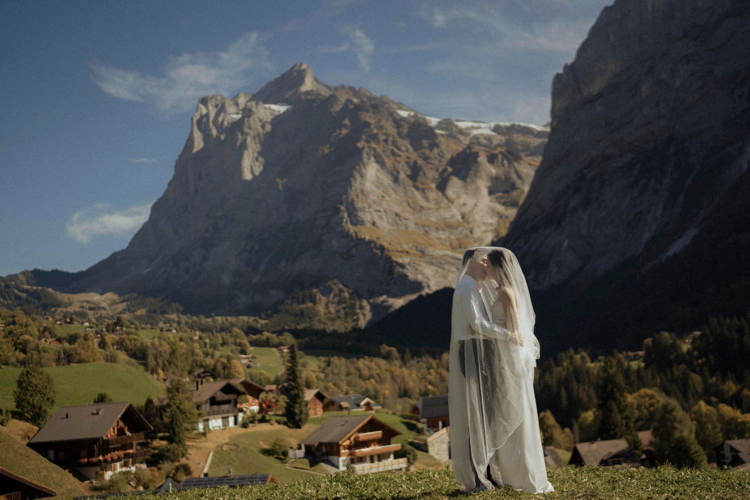 Wedding couple standing on mountain meadow with dramatic Alpine peaks in background.