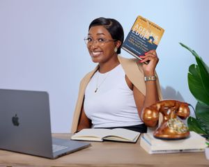 A lady seating on a desk with a laptop holding a real estate book