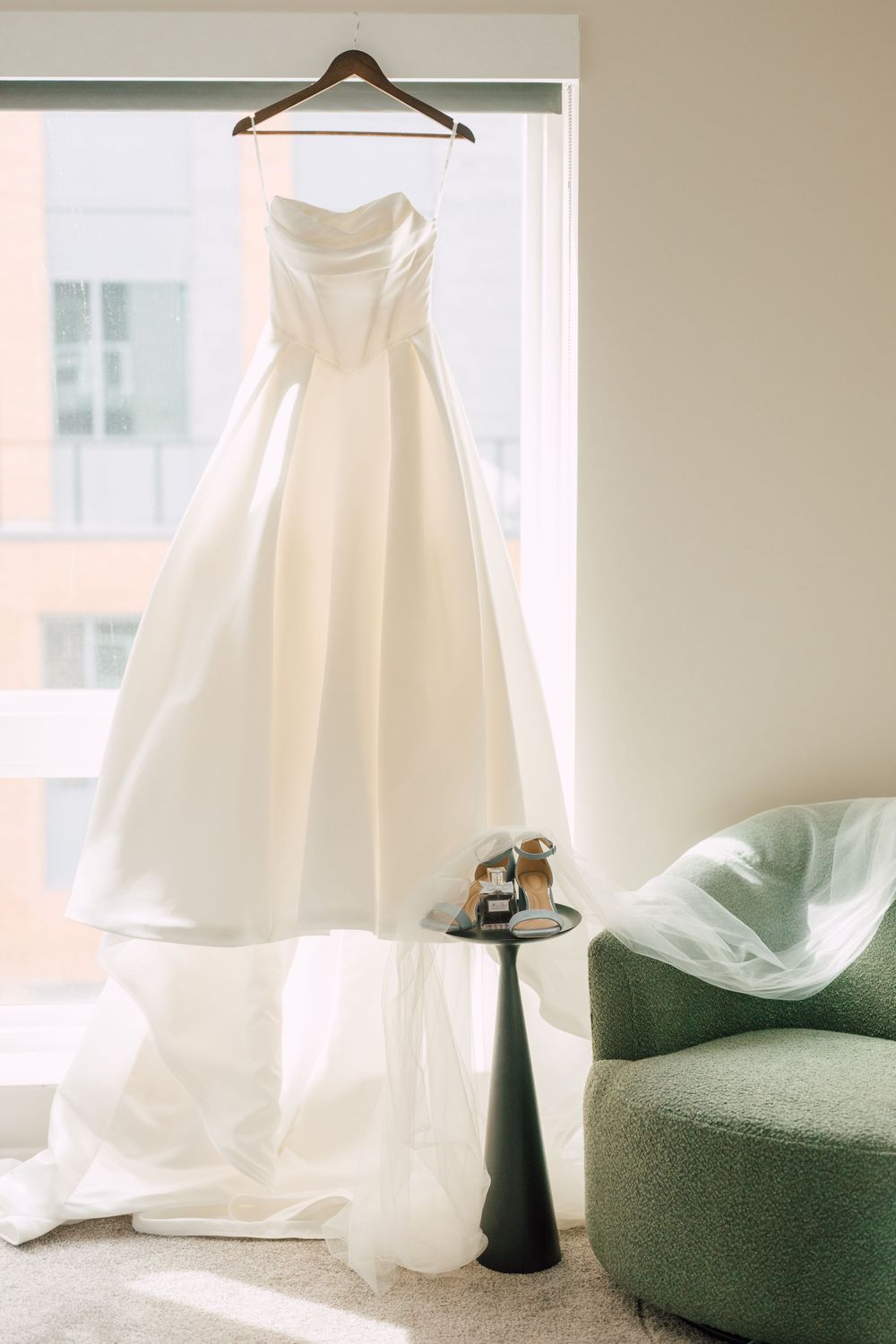 Elegant strapless white wedding gown hanging by window with green accent chair and bridal shoes on side table.