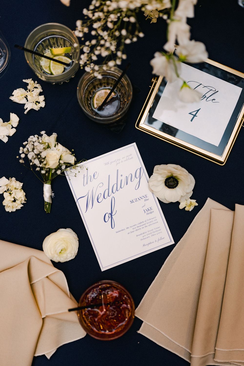 Elegant wedding flat lay with navy tablecloth, floral invitation, table number 4, white flowers, and glassware.