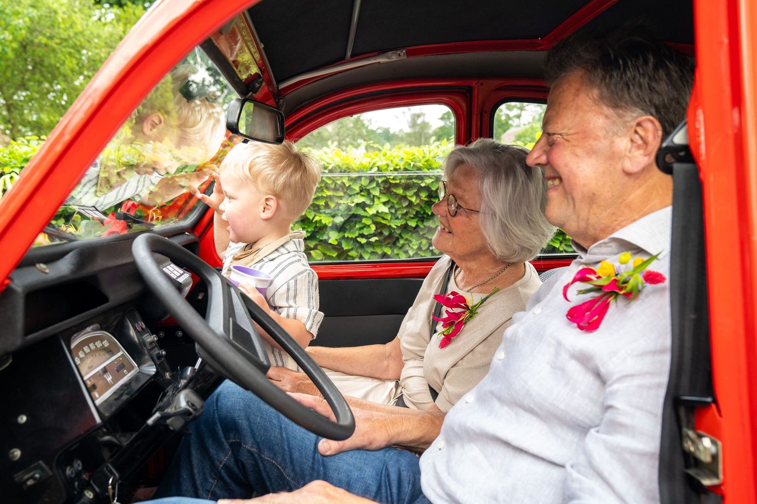 Grootouders en kleinzoon lachen samen in een vrolijke rode oldtimer op een feestdag.