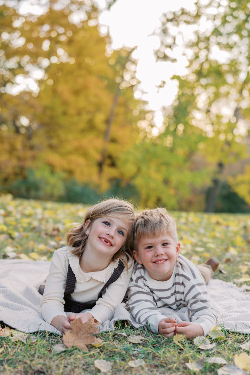 Two people lie on their stomachs on a white blanket surrounded by yellow autumn leaves.