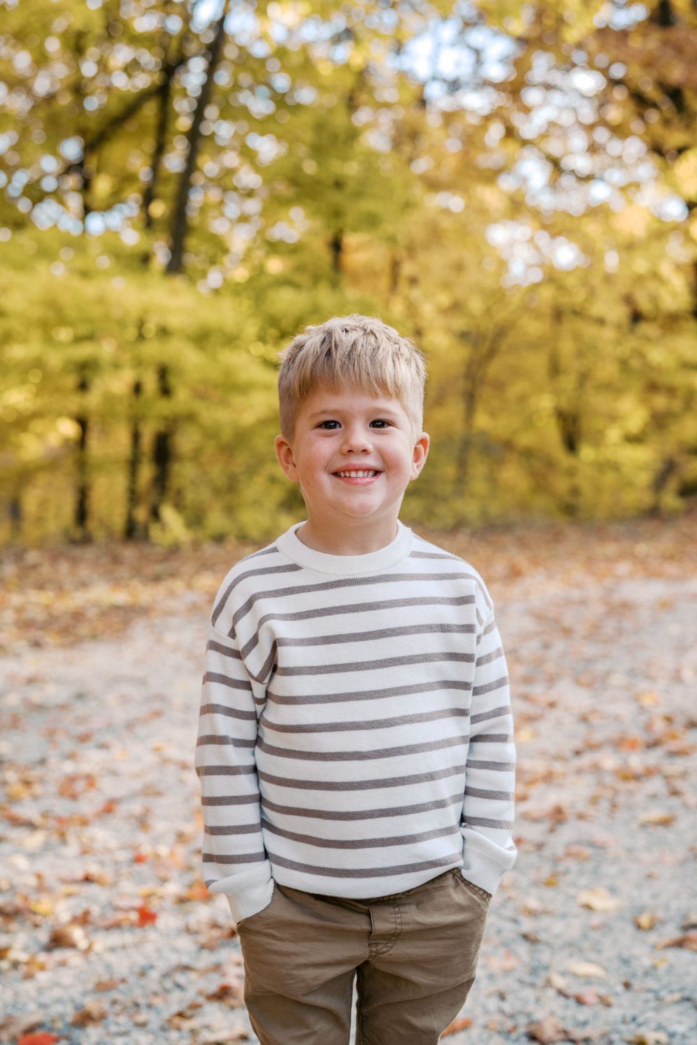 Child in striped sweater smiling during autumn photoshoot with yellow trees in background.