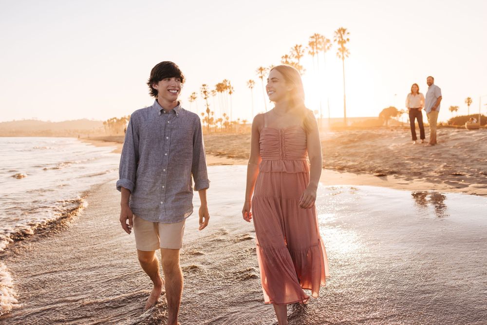 A couple strolls along a beach at sunset with palm trees silhouetted against the golden sky.