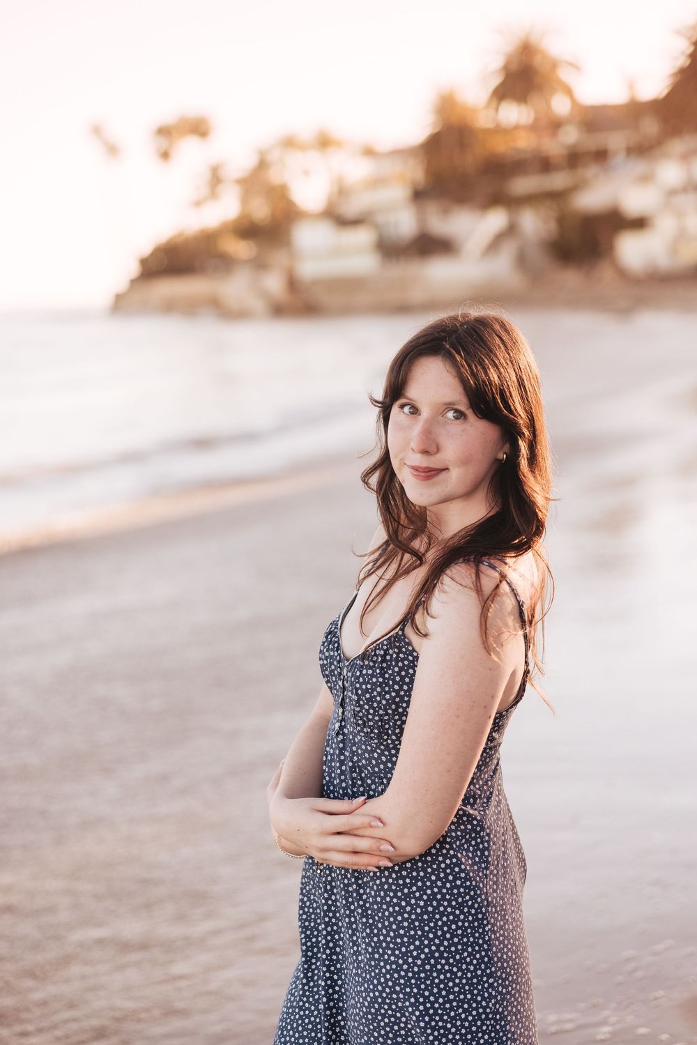 Young woman in polka dot sundress poses on a sandy beach at golden hour sunset with ocean waves behind her.