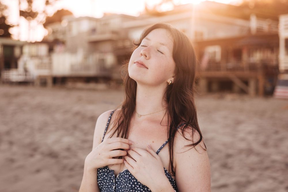 Young woman with dark hair enjoys golden sunset light on a sandy beach, hands clasped to chest, eyes closed.