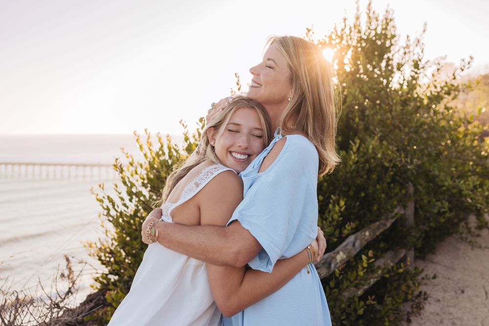 Two friends in white and blue dresses embrace while laughing together at sunset by the beach.