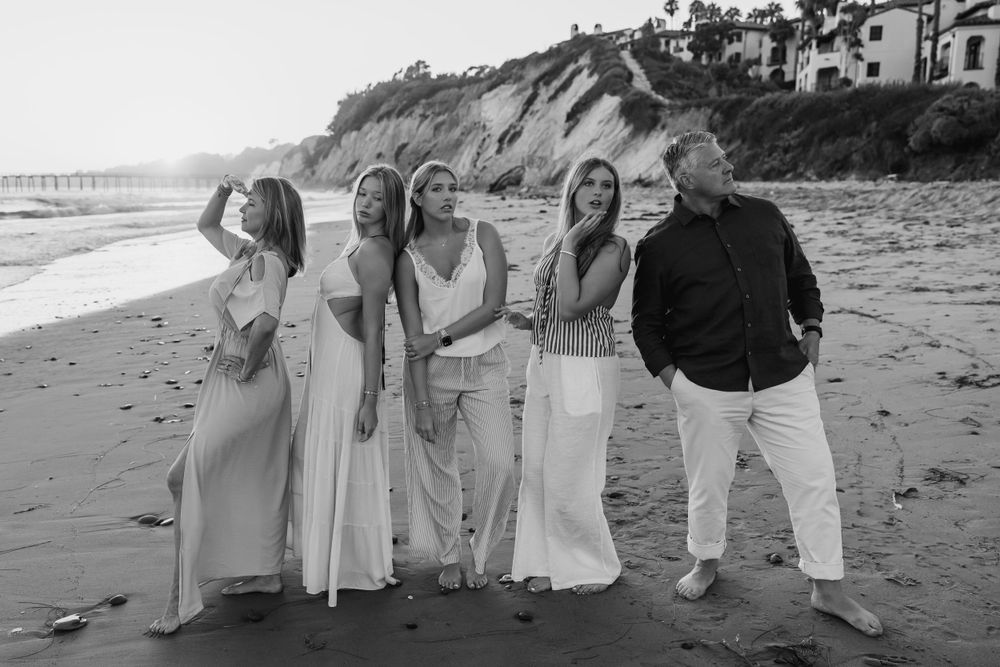A black and white photo showing a group of people in light-colored outfits standing together on a beach with cliffs behind them.