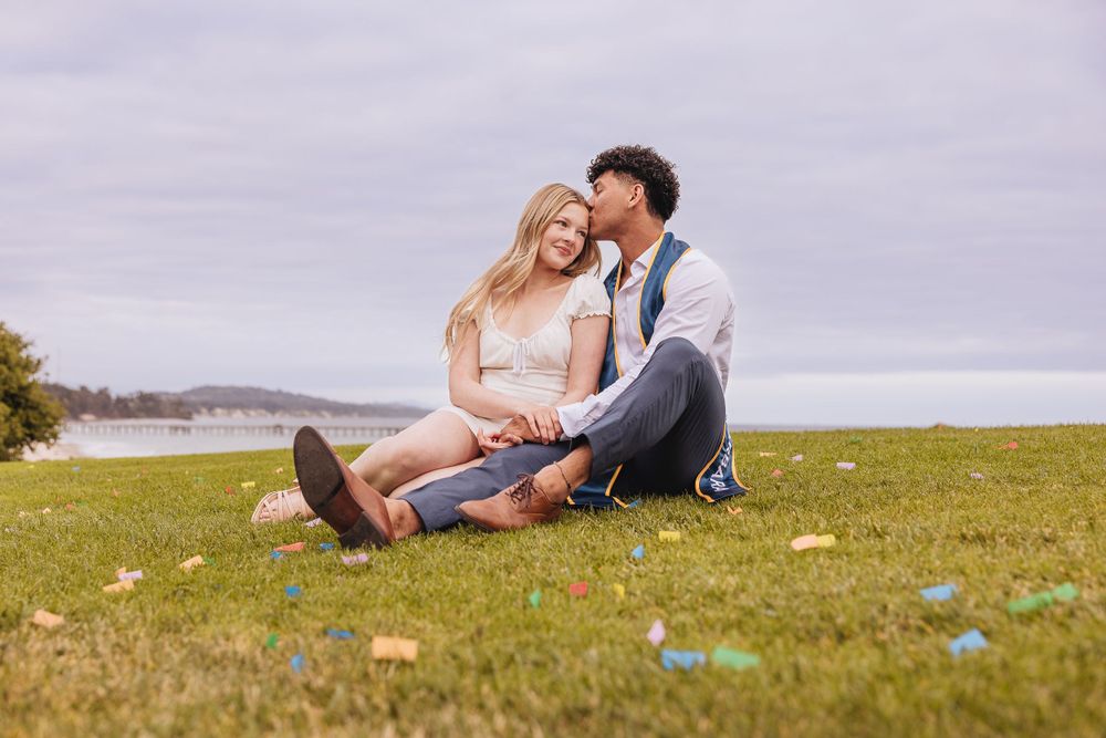 Two people sitting on green grass by a waterfront on a cloudy day with colorful Easter eggs scattered around.