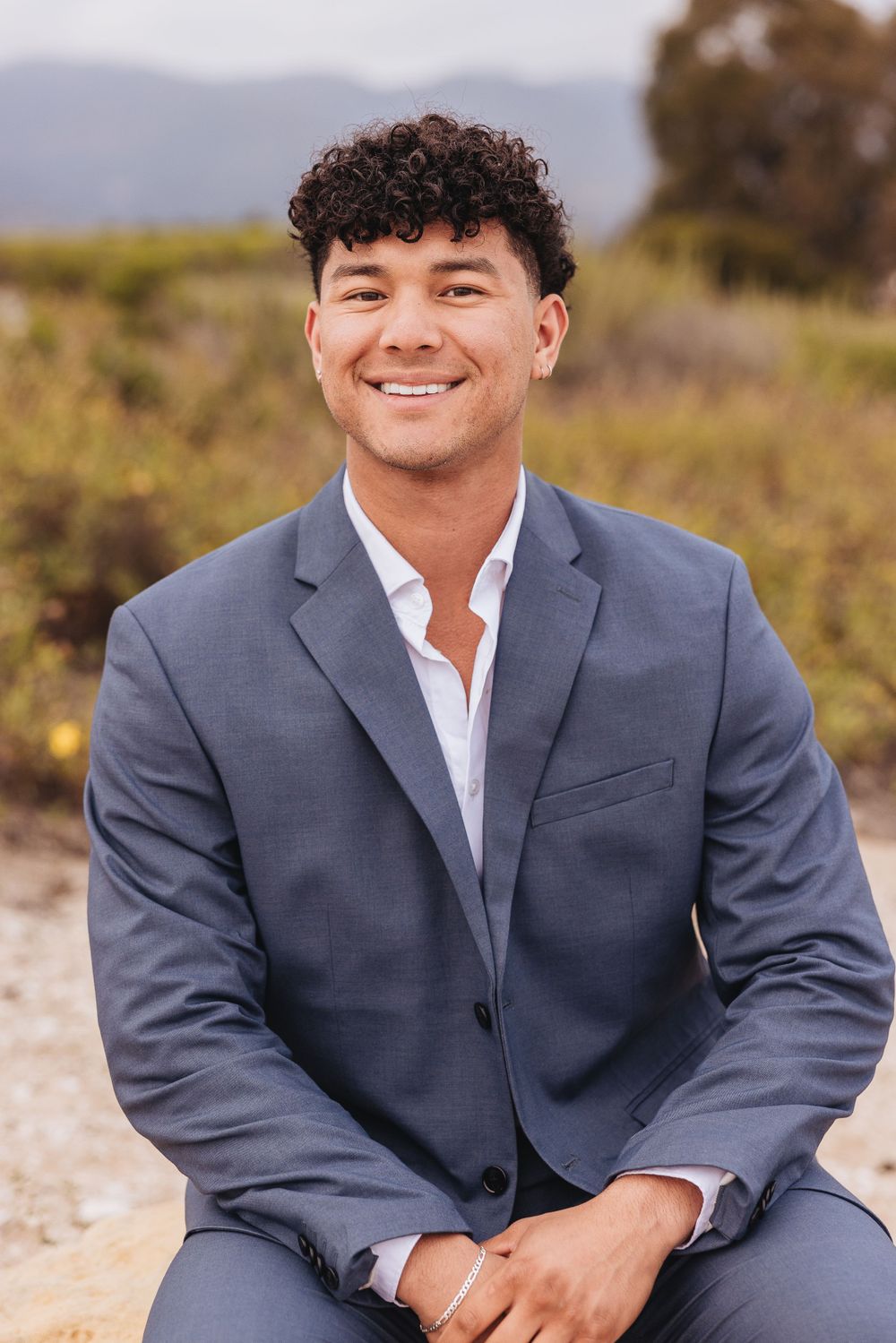 Professional portrait of a person in a gray suit and white shirt outdoors with blurred natural background.