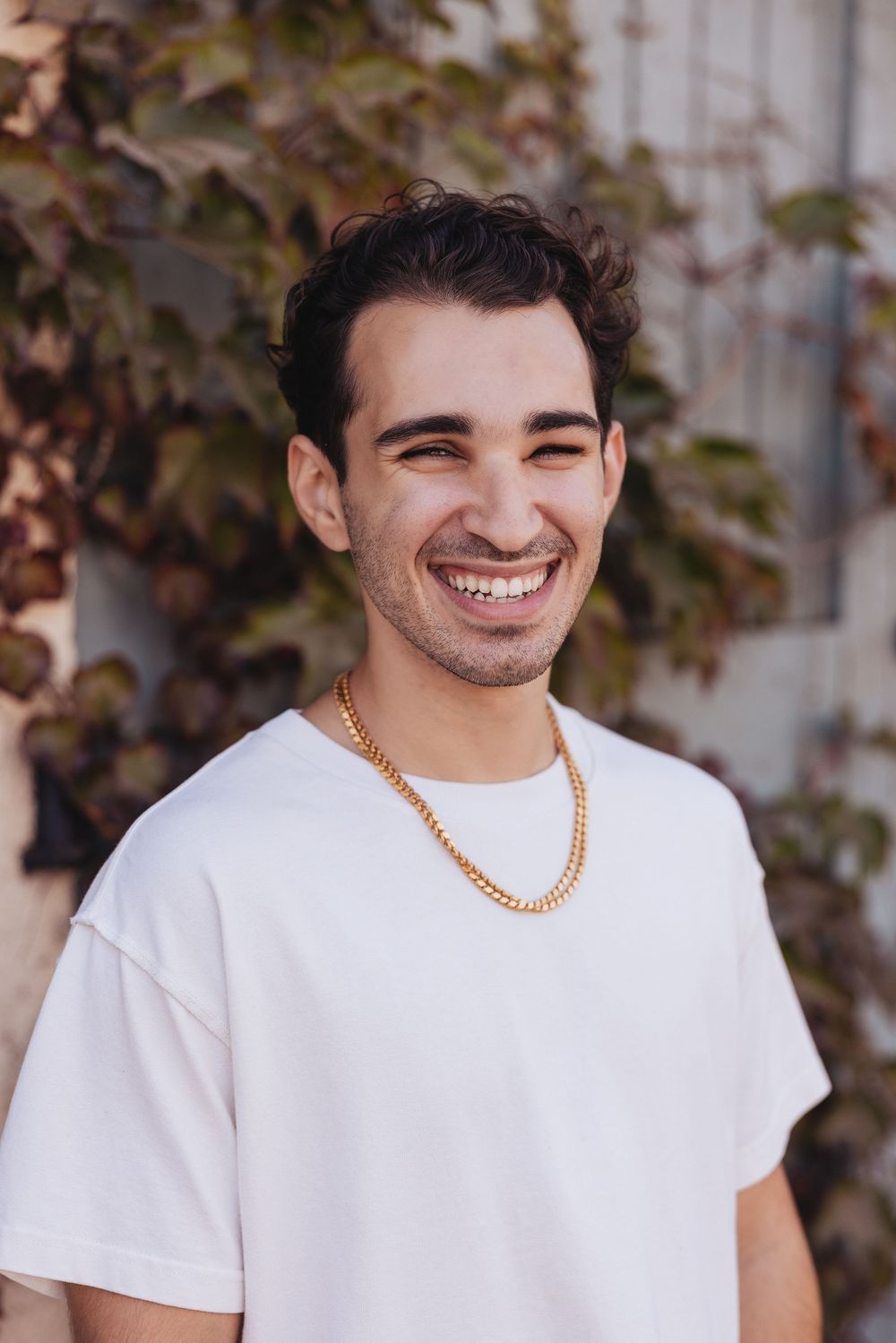 Smiling young man in white t-shirt and gold chain necklace standing outdoors near ivy-covered wall.