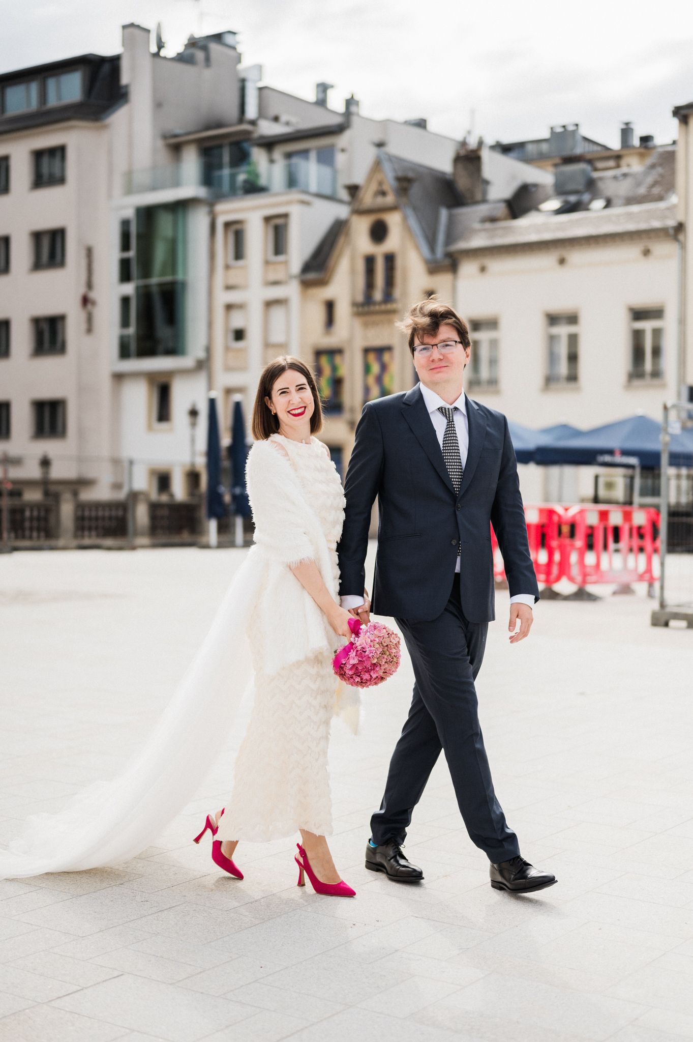 A wedding couple walks down a European street, with the bride in white gown and red heels holding a pink bouquet.