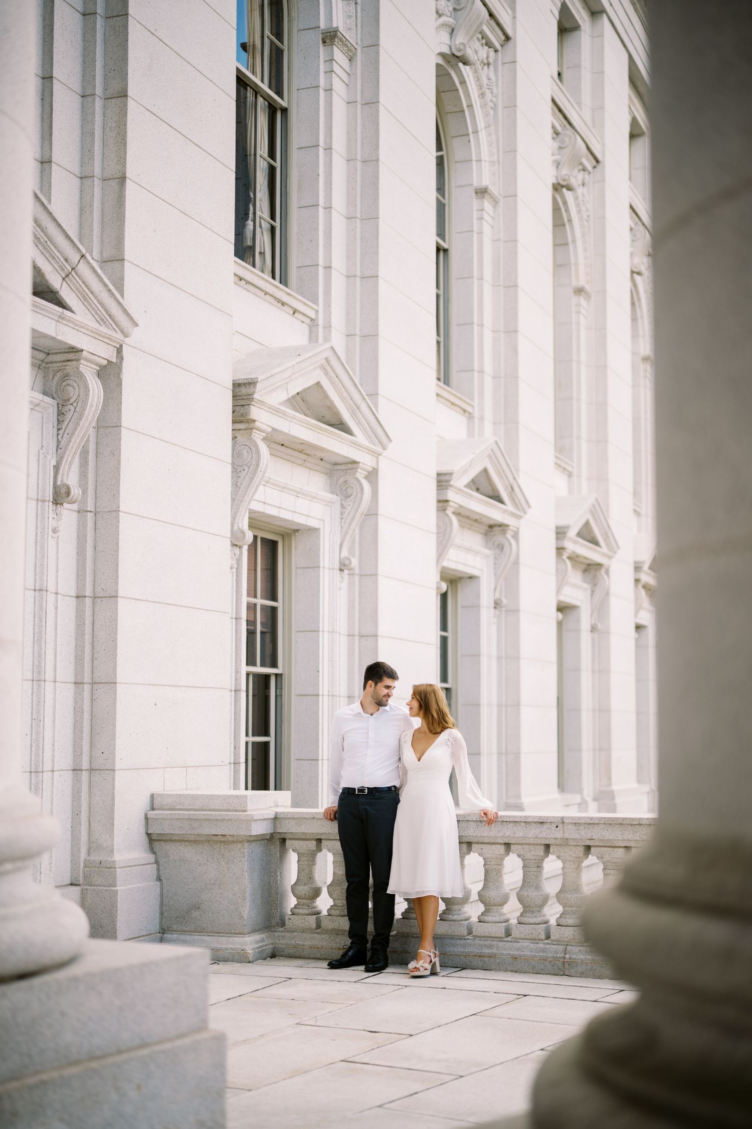 A romantic couple in white and black attire stands together against a classical white stone building with ornate columns.