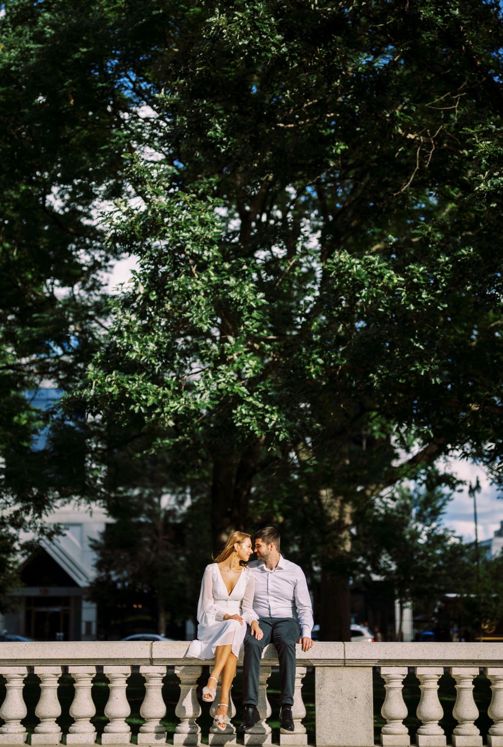 Couple sitting close together on stone balustrade under large leafy tree on sunny summer day.