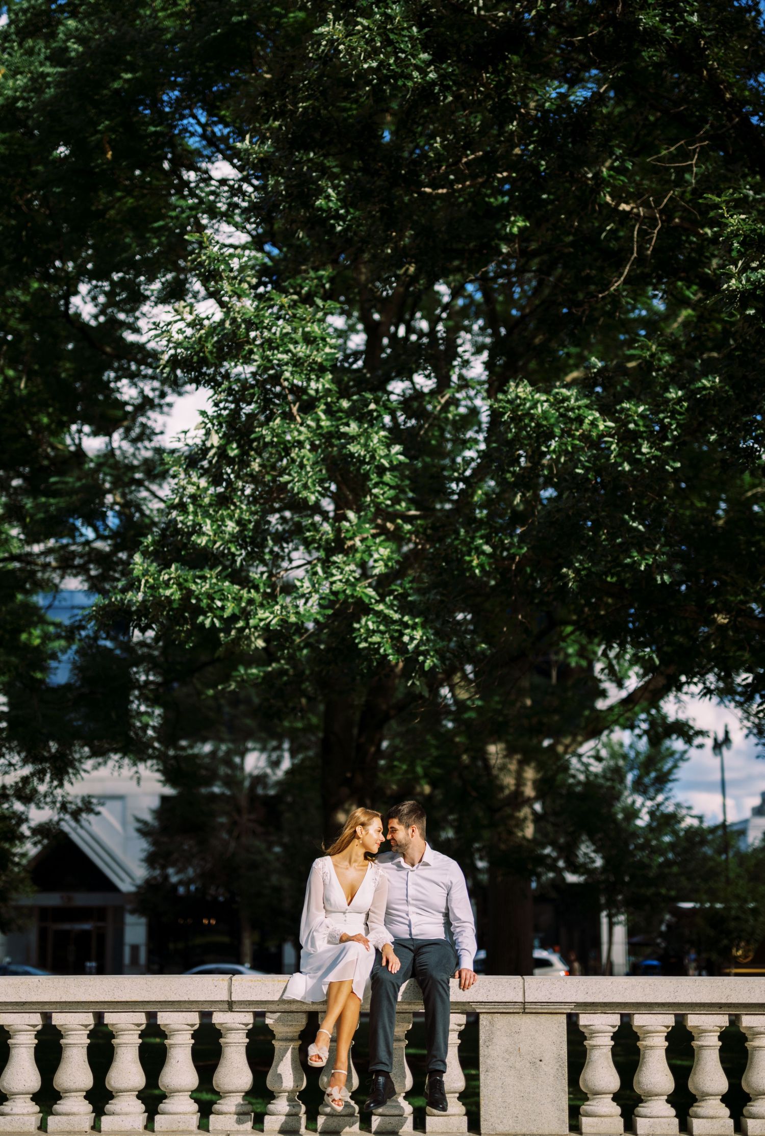 Couple sitting close together on stone balustrade under large leafy tree on sunny summer day.