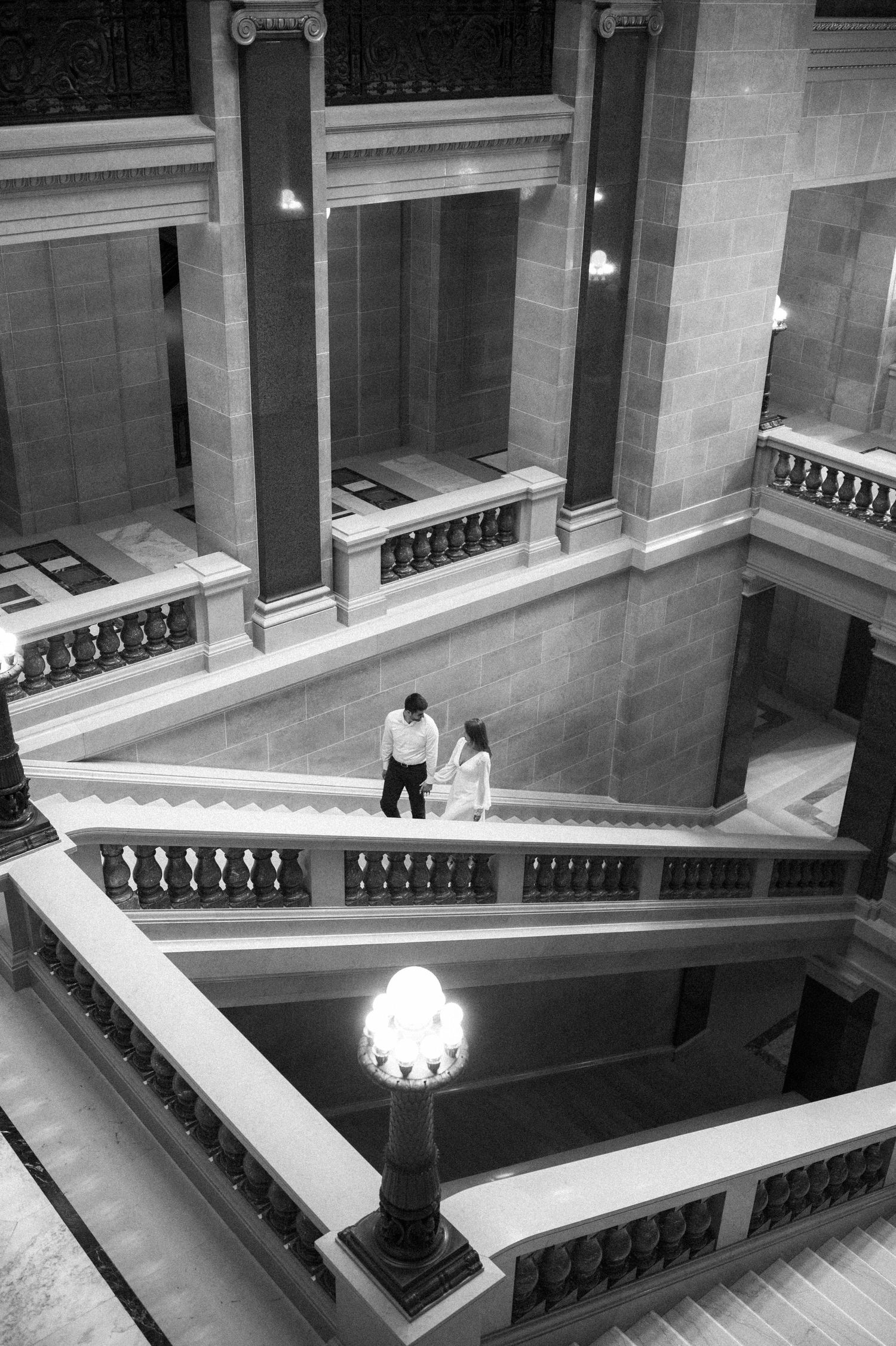 Two people descending an elegant marble staircase in a grand building with ornate architectural details and vintage lighting.