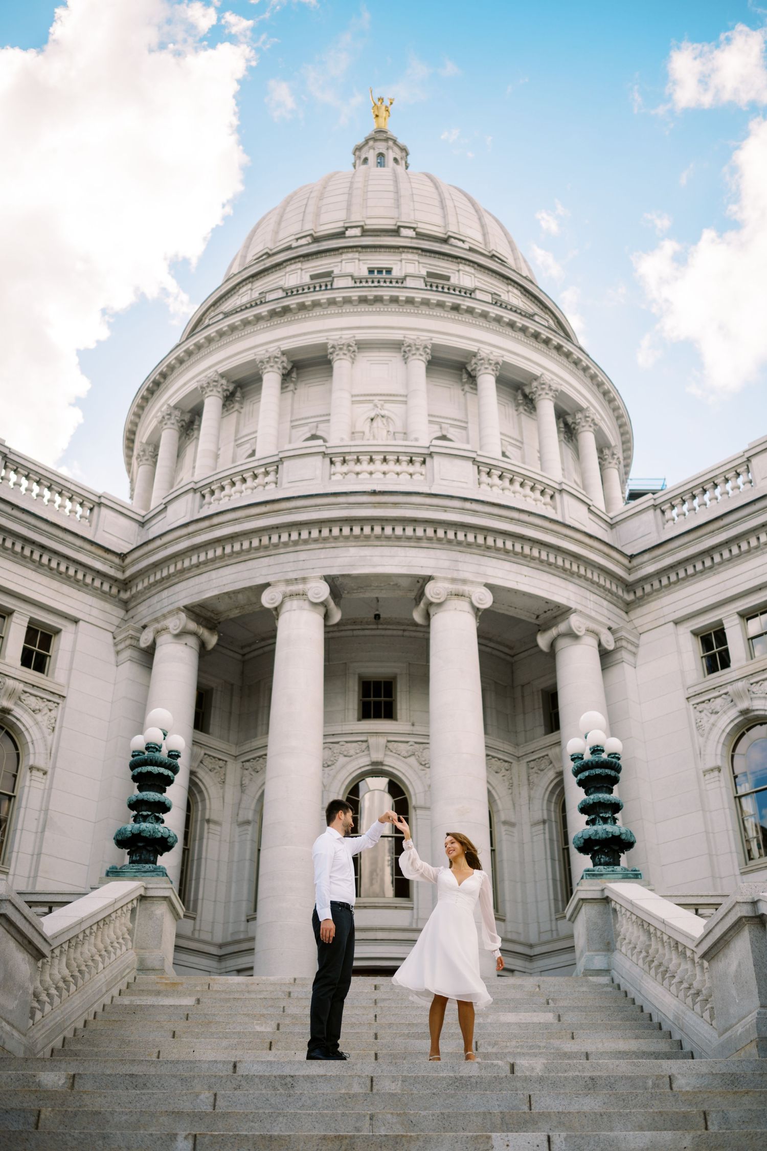 Two people stand on the grand steps of the Wisconsin State Capitol building with its iconic dome and neoclassical architecture.