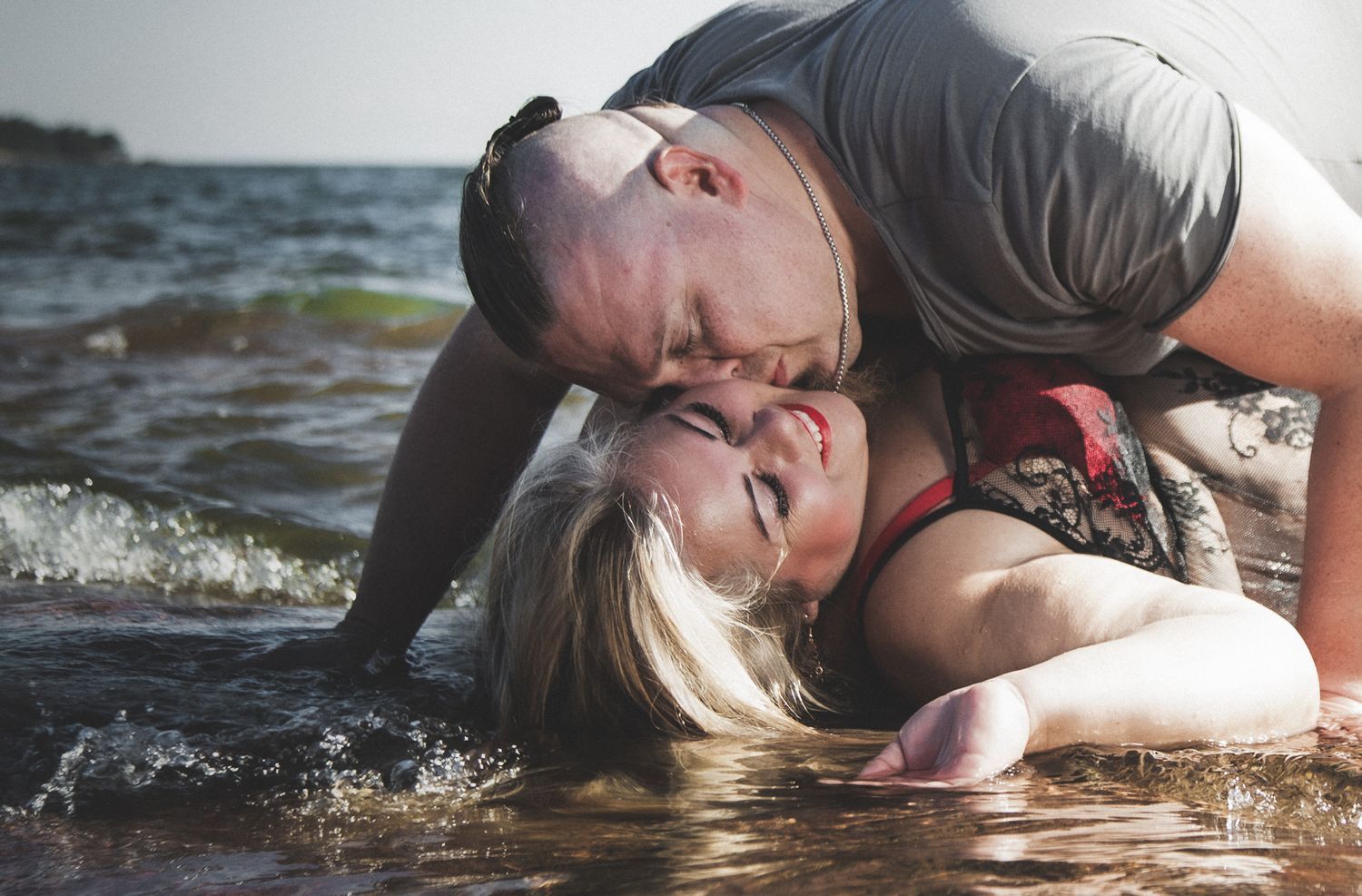 Couple playing in shallow ocean water with waves splashing around them during a romantic photoshoot.