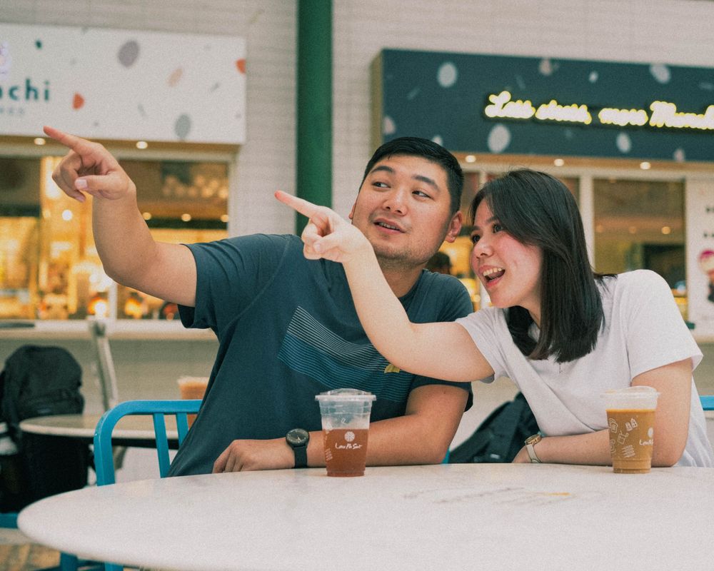 Playful moment between couple pointing and smiling while enjoying beverages at outdoor cafe.