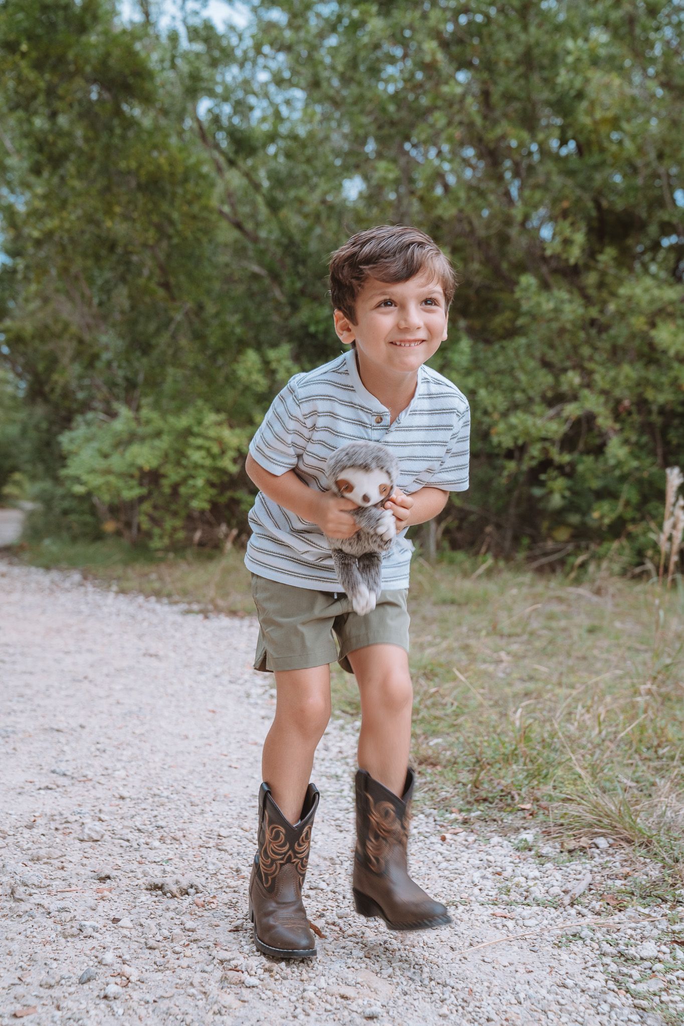 A child wearing cowboy boots and shorts plays outdoors on a gravel path surrounded by green foliage.