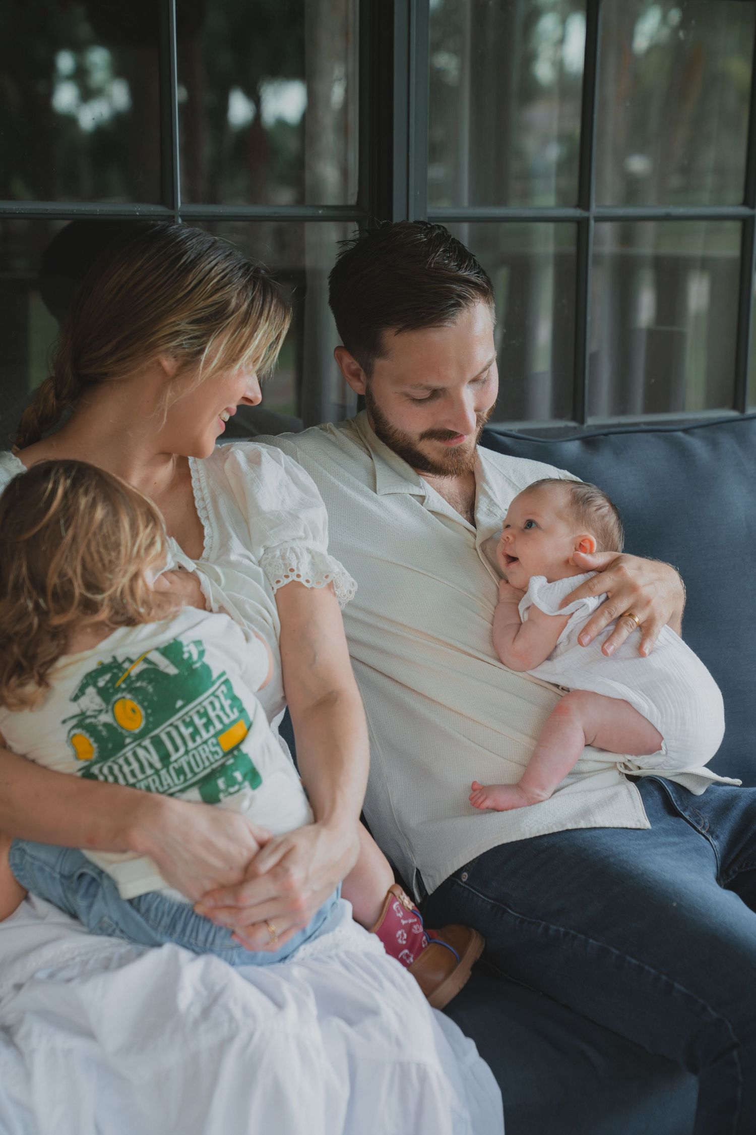 Young family sitting on sofa, mother holding toddler in John Deere shirt, father cradling newborn baby.