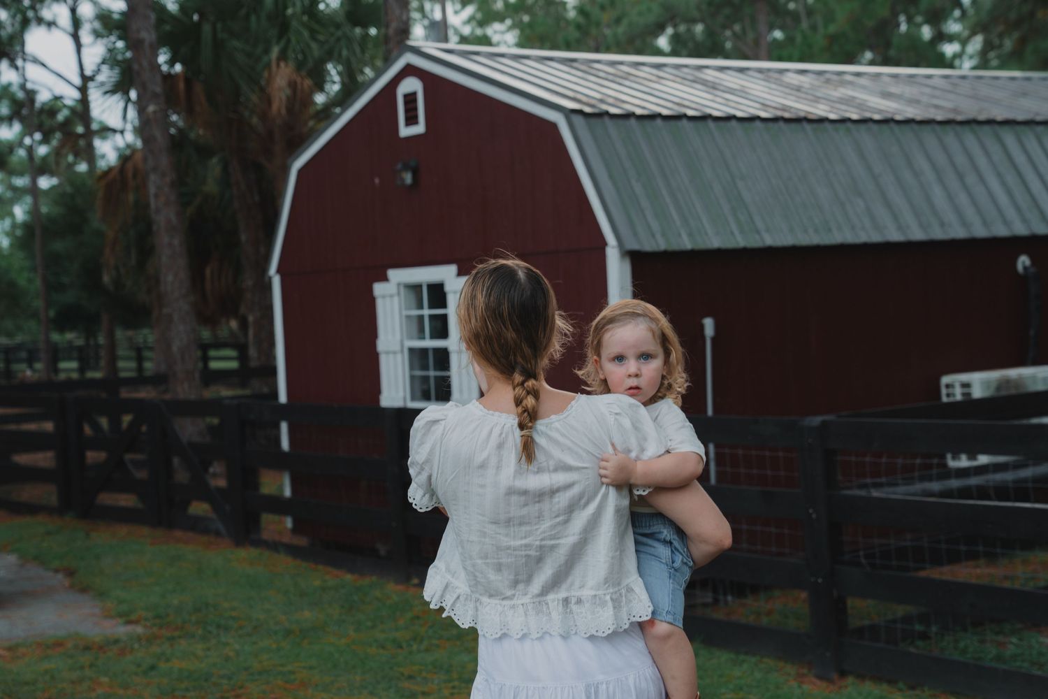 A mother carries her young daughter near a red barn on a farm, both dressed in casual summer clothing.