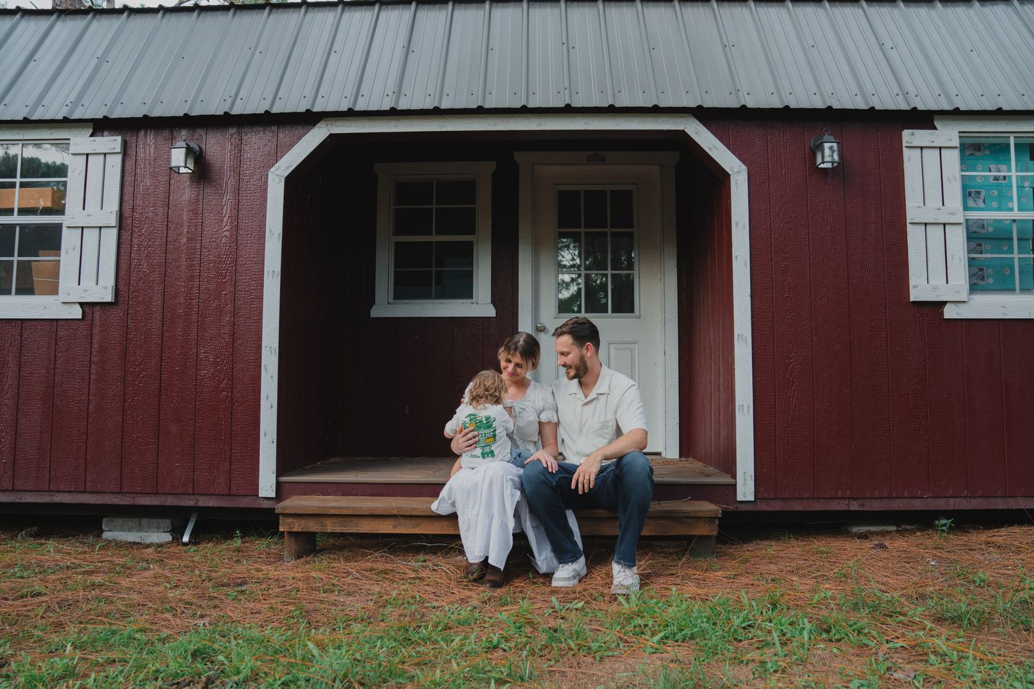 A couple sits on the steps of a red barn-style cabin, sharing a tender moment with their newborn baby outdoors.
