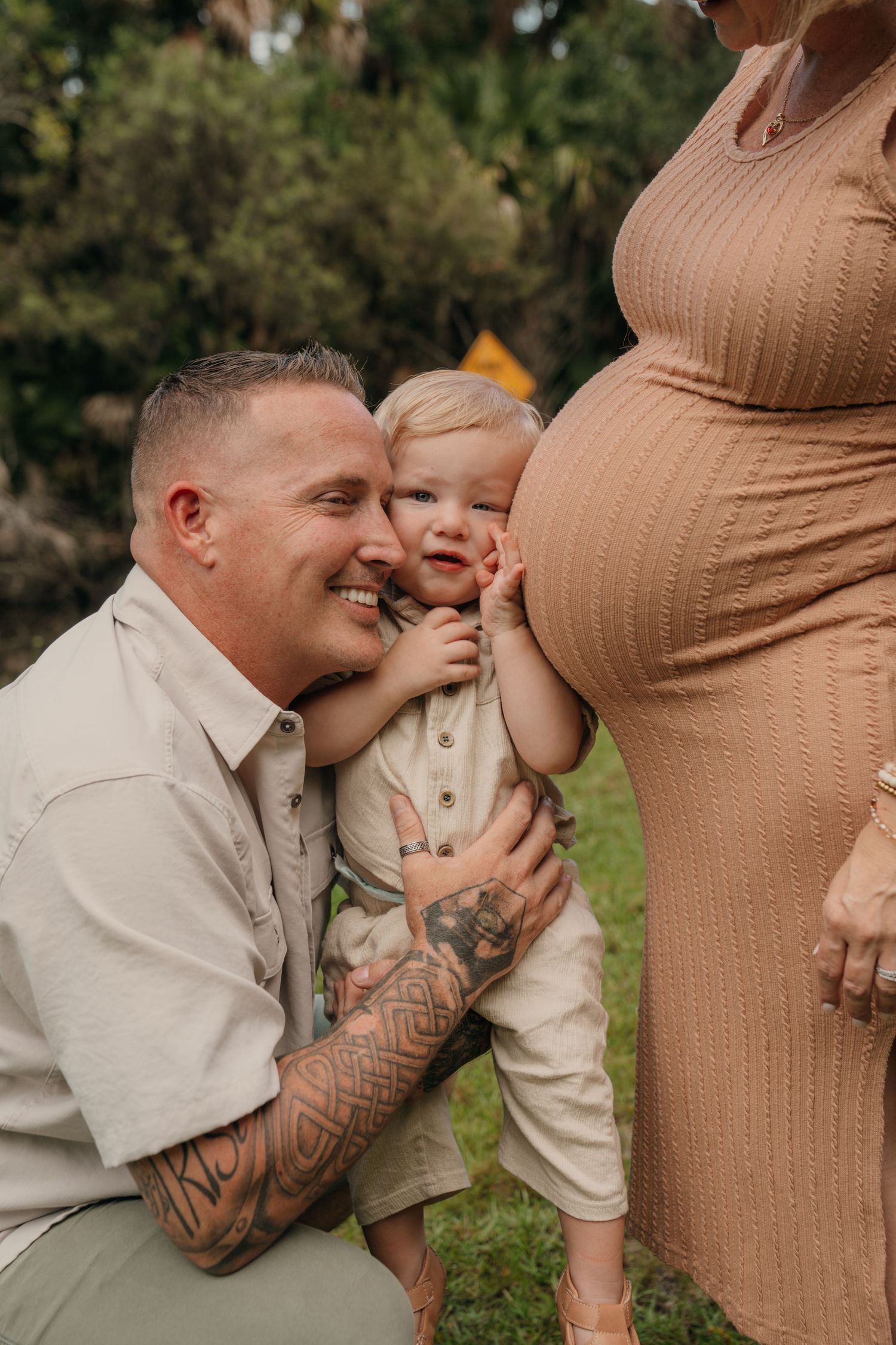 A tattooed father kneels beside his toddler touching a pregnant woman's baby bump outdoors.
