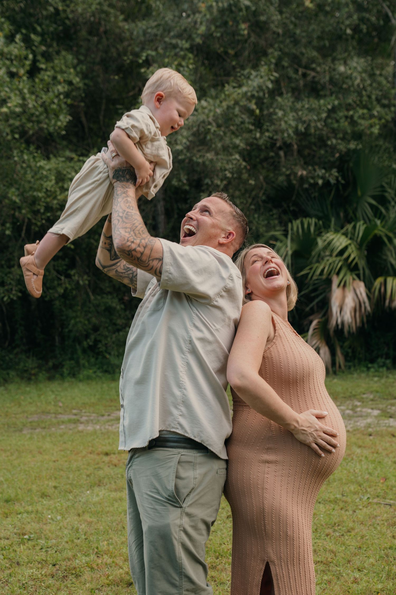 Happy family outdoors, man lifting toddler while laughing pregnant woman in tan dress leans against him.