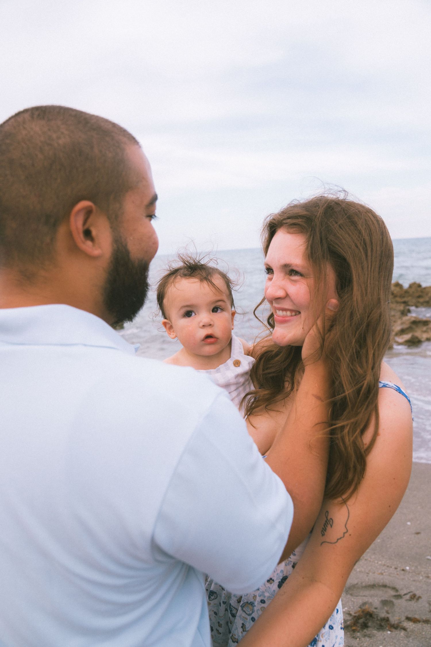 A couple smiles at each other on a rocky beach while holding their baby girl in a heartwarming family portrait.