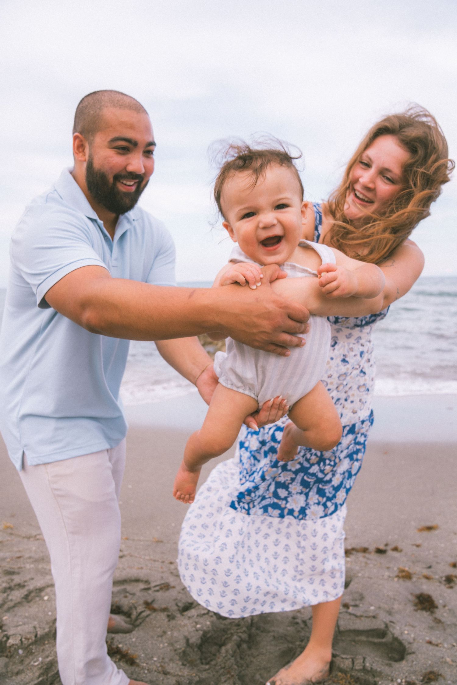 Happy family with laughing baby enjoying a fun day at the beach, parents smiling and holding child on sandy shore.
