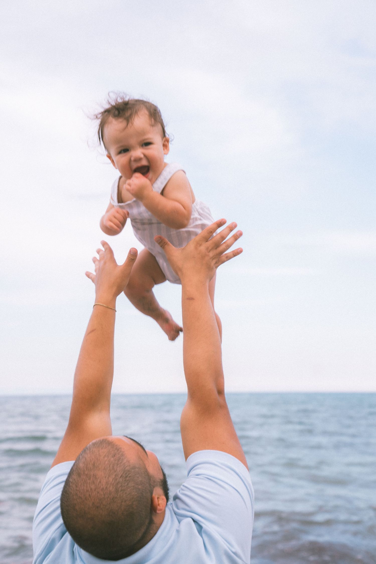 Father joyfully lifting laughing baby in the air at the beach with ocean in the background.